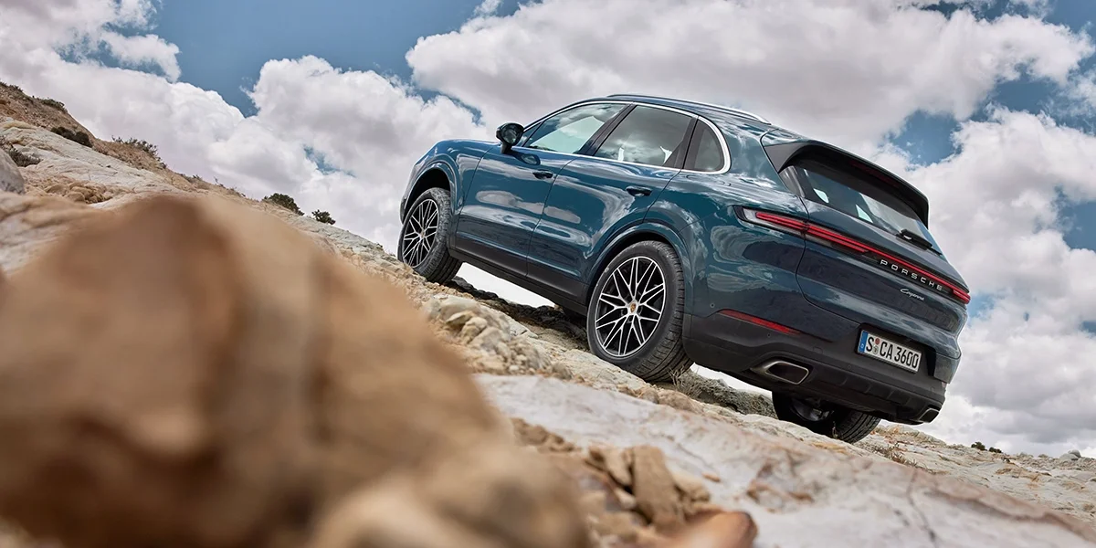 Rear view of 2026 Porsche Cayenne climbing a rocky incline under a partly cloudy sky.