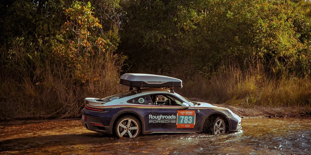 Porsche 911 Dakar driving through water with roof box and rally decals in an off-road setting
